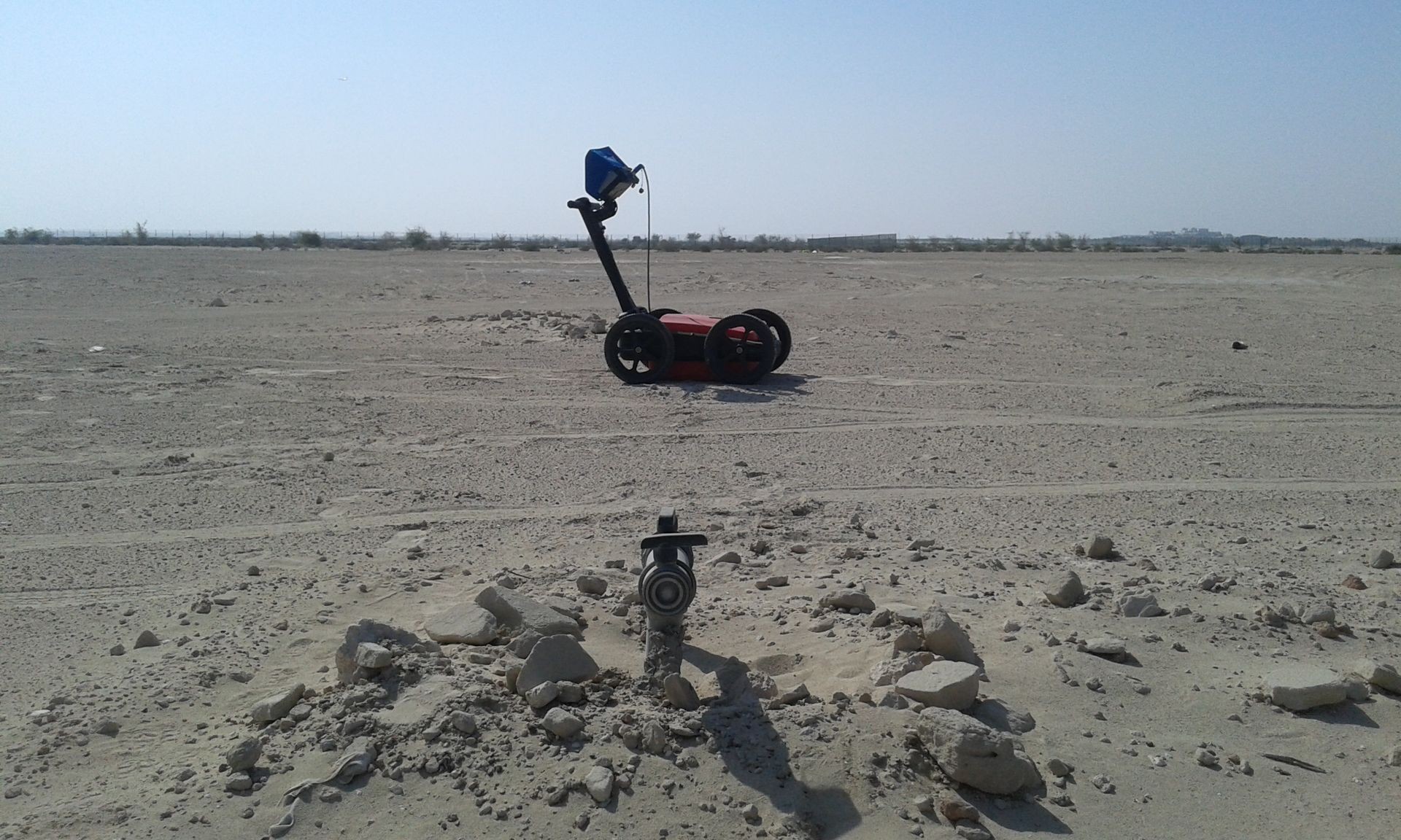 Robot with wheels on a sandy terrain, distant horizon with sparse greenery.