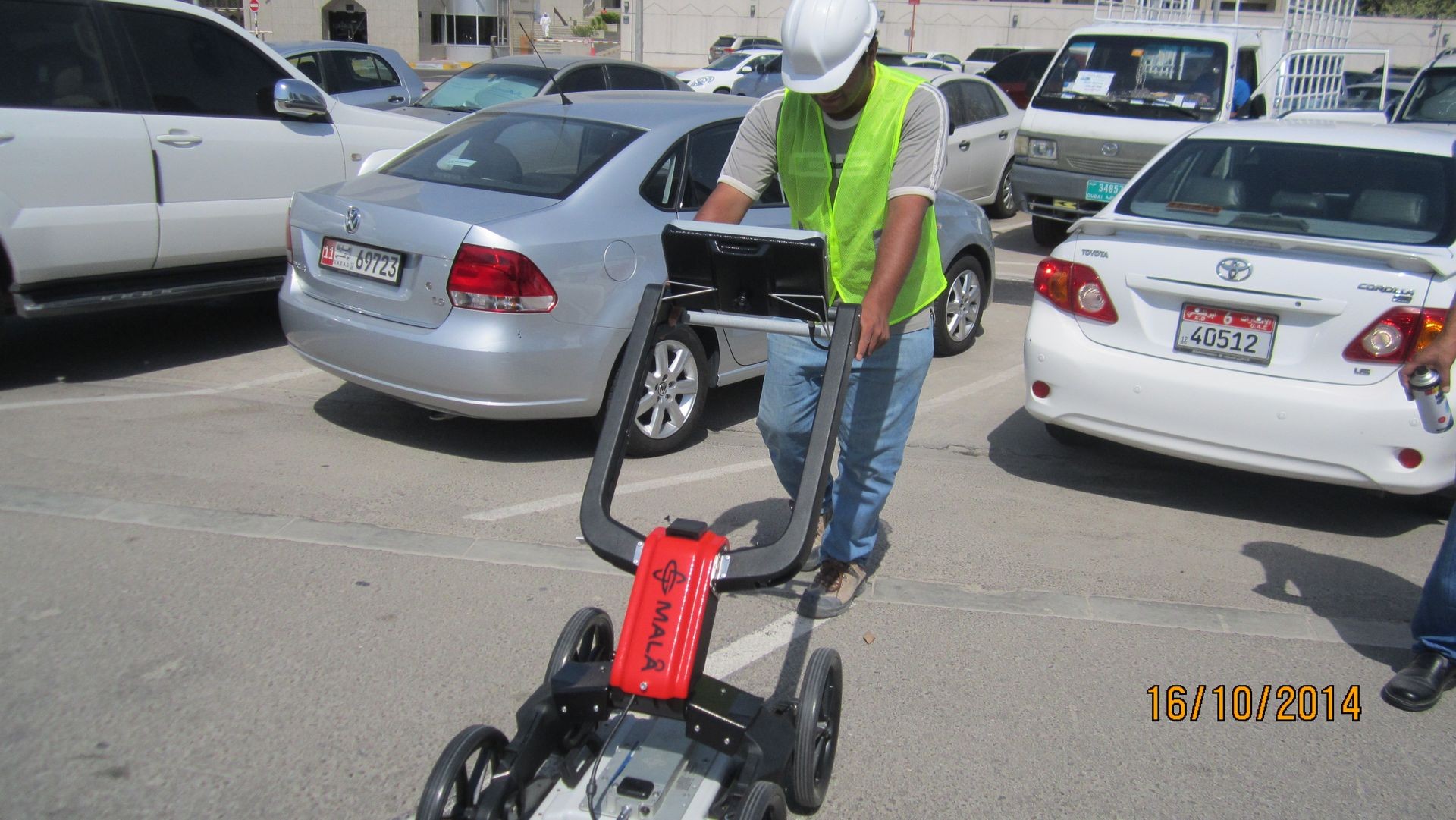 Construction worker in safety gear using a ground-penetrating radar device in a parking lot.